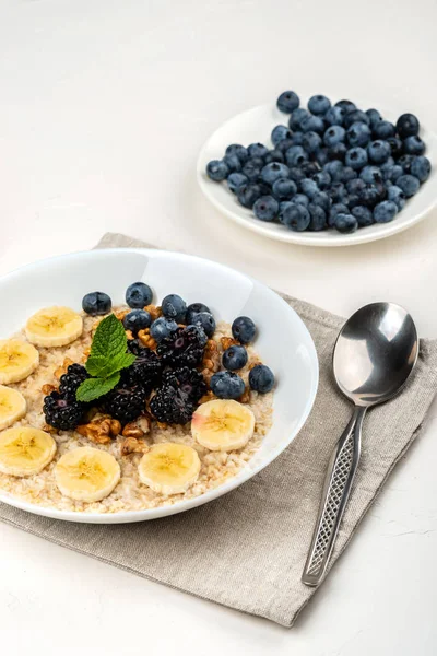 Овсяная каша с бананами, грецкими орехами, ежевикой, черникой, медом и мятой в белой миске на белом фоне. Здоровый завтрак и домашняя диета . — стоковое фото