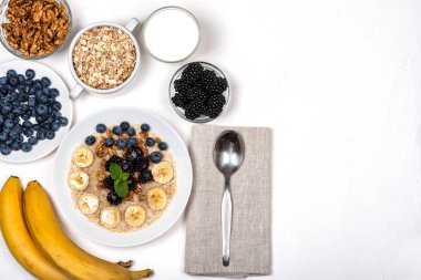 Muz, ceviz, böğürtlen, yaban mersini, bal ve nane ile yulaf lapası beyaz bir arka plan üzerinde beyaz bir kapta. Sağlıklı kahvaltı ve ev yapımı diyet gıda. Düz döşeme stilinde üst görünüm.