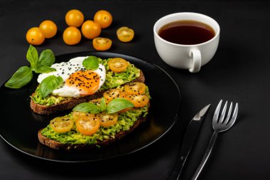 Açık sandviç avokado guacamole, sarı kiraz domates, kızarmış yumurta ve fesleğen siyah arka plan üzerinde siyah bir tabak üzerinde. Sağlıklı yiyecek veya atıştırmalık