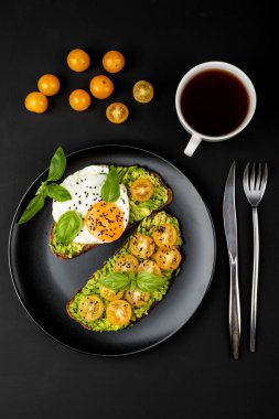 Açık sandviç avokado guacamole, sarı kiraz domates, kızarmış yumurta ve fesleğen siyah arka plan üzerinde siyah bir tabak üzerinde. Üstte görüntü.