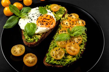 Açık sandviç avokado guacamole, sarı kiraz domates, kızarmış yumurta ve fesleğen siyah arka plan üzerinde siyah bir tabak üzerinde. Sağlıklı yiyecek veya atıştırmalık