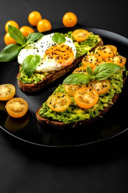Açık sandviç avokado guacamole, sarı kiraz domates, kızarmış yumurta ve fesleğen siyah arka plan üzerinde siyah bir tabak üzerinde. Sağlıklı yiyecek veya atıştırmalık