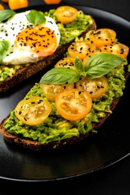 Açık sandviç avokado guacamole, sarı kiraz domates, kızarmış yumurta ve fesleğen siyah arka plan üzerinde siyah bir tabak üzerinde. Sağlıklı yiyecek veya atıştırmalık