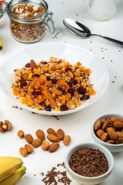 Sağlıklı kahvaltı. Beyaz arka planda beyaz bir kasede süt, kurutulmuş kayısı, kızılcık, badem ve keten tohumlu müsli. Temiz yemek, diyet, detoks, vejetaryen yiyecekler.