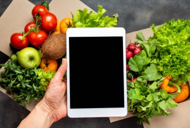 Yemek teslimatı. Kadın eli, tablet bilgisayarı taze organik ürünlerle dolu kutuların üzerinde boş ekranla tutuyor..