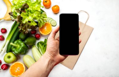 Sipariş, internete yemek teslimatı. Kadın eli, organik sebzeler üzerinden mesajınız için boş ekranlı akıllı telefonu tutuyor. Yakın plan.