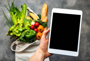 Tablet bilgisayarı, içi sağlıklı yiyeceklerle dolu alışveriş çantasının üzerinde boş ekranlı kadın eli. İnternetten yemek siparişi vermek.