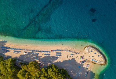 Podgora 'daki Makarska Riviera sahilinin insansız hava aracı görüntüsü düzenli güneş yatağı hatları sakin sabah denizi ve koruyucu kaya iskelesi.