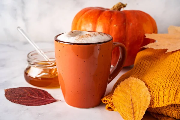 Close up of orange cup with coffee or pumpkin spice latte, autumn leaves, pumpkin, and honey jar on white background