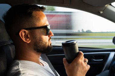 Güneş gözlüklü, kendine güveni tam sakallı bir adam otoyolda arabasını sürerken tekrar kullanılabilir bir kahve bardağı tutuyor..