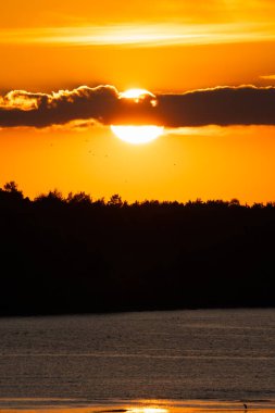 Güneşin bulutların arkasında, siluetli bir orman ve Estonya manzaralı sakin bir göl üzerinde dramatik ateşli turuncu günbatımı..