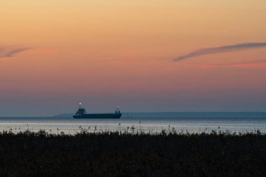 Kargo gemisi alacakaranlıkta sakin Baltık Denizi 'nde yol alırken, soluk kesici bir gökyüzü turuncu ve mor yumuşak renkler sergiliyor, sükuneti yansıtıyor..