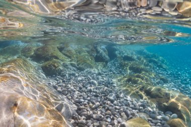 Kristal berrak su, su yüzeyindeki parıldayan güneş ışığıyla aydınlatılan taş ve çakıl taşlarından oluşan güzel bir deniz yatağını ortaya çıkarır..