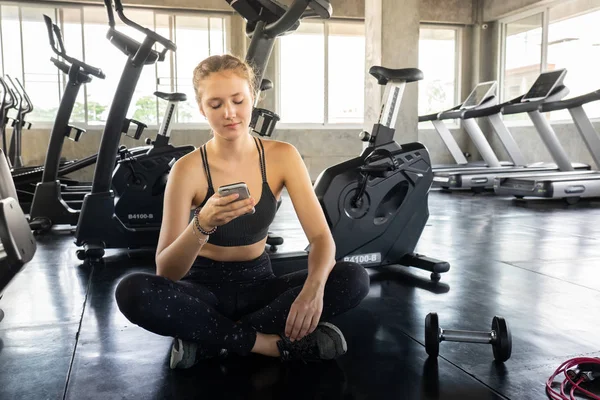 Fitness salonunda Smartphone kullanan etkin kız. Rahatlatıcı ve bir cep telefonu kullanarak müzik salonunda genç gülümseyen kadın