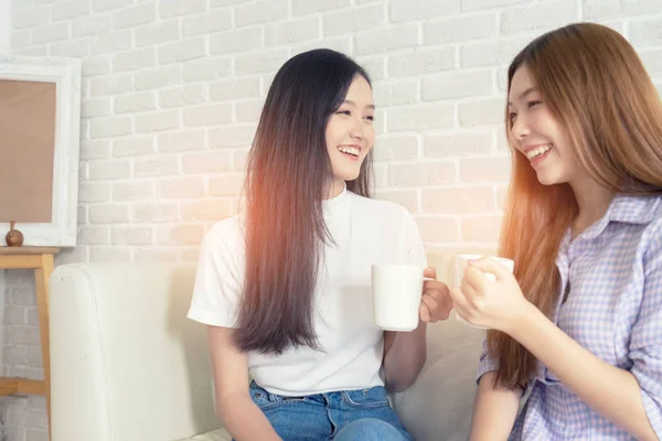Happy female friends holding coffee mugs while discussing at table in living room relax time.Asian  beauty woman smile and sititing sofa .
