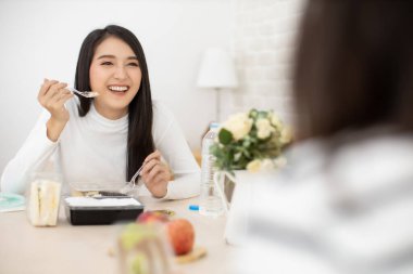 Genç Asyalı bir kadın restoranda ya da evde ayrı oturuyor. Masa başında yemek yiyor. Enfeksiyonu Coronavirus covid-19, yeni normal restoran ve sosyal uzaklık kavramından koruyor..