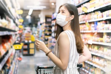 Yüz maskesi takan Asyalı kadın akşam yemeği pazarında alışveriş arabası satıyor. Coronavirus krizi sırasında ya da covid19.mutlu yaşam tarzı ya da alışveriş anlayışı sırasında raftan bir şeyler almak isteyen bir kız..