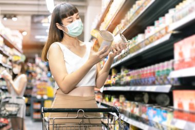 Yüz maskesi takan Asyalı kadın akşam yemeği pazarında alışveriş arabası satıyor. Coronavirus krizi sırasında ya da covid19.mutlu yaşam tarzı ya da alışveriş anlayışı sırasında raftan bir şeyler almak isteyen bir kız..