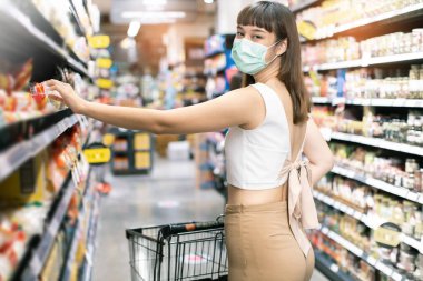 Yüz maskesi takan Asyalı kadın akşam yemeği pazarında alışveriş arabası satıyor. Coronavirus krizi sırasında ya da covid19.mutlu yaşam tarzı ya da alışveriş anlayışı sırasında raftan bir şeyler almak isteyen bir kız..