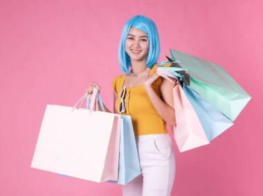 Renkli alışveriş çantalarıyla oldukça heyecanlı Asyalı bir kızın portresi pembe arka planda soyutlanmış görünüyor. Alışveriş torbaları taşıyan alışveriş kolik bir kadın yaz indirimi konsepti için. Genç mavi saçlı kız. .