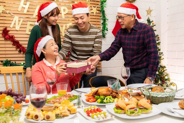 Thanksgiving or christmas Celebration Asian  Family Dinner Concept.Asian happy family celebrating Christmas with Grandparents while sitting over dining chair and wearing Santa Hat and with  gift box.
