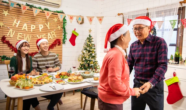 Thanksgiving or christmas Celebration Asian  Family Dinner Concept.Asian happy family celebrating Christmas with Grandparents while sitting over dining chair and wearing Santa Hat and with  gift box.