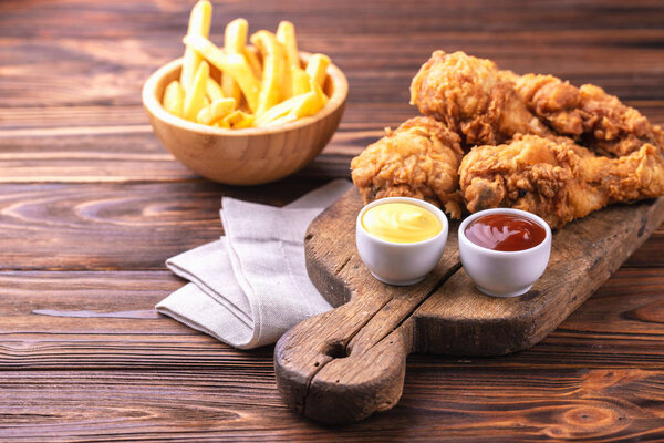 Жареные хрустящие и пряные куриные ножки и картофель фри с соусами. Концепция быстрого и нездорового питания. Вид сверху, копипространство
