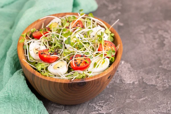 Light salad with radish sprouts, quail eggs, cherry tomatoes and sesame in wooden bowl on dark concrete background. Copy space
