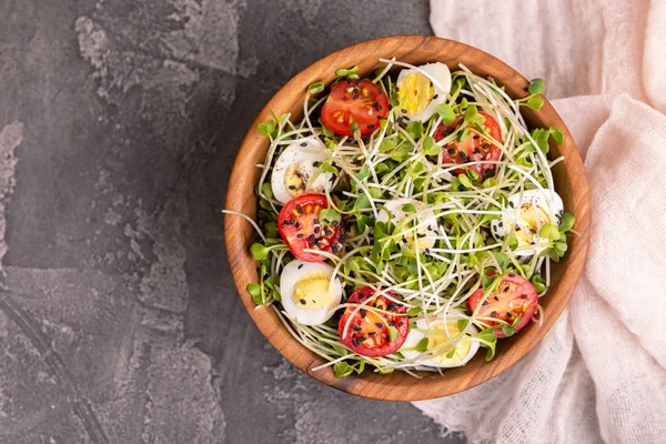 Light salad with radish sprouts, quail eggs, cherry tomatoes and sesame in wooden bowl on dark concrete background. Copy space. Top view