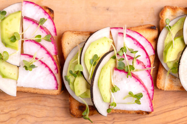 Tasty wheat toasts with radish, avocado and sprouts on wooden cutting board. Dark concrete background. Simple healthy breakfast. Copy space. Top view