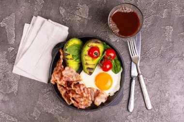 Izgara domuz pastırması ve avokado, ıspanak ve kiraz domates dökme demir tavada kızarmış yumurta. Gri beton zemin. Üstten görünüm. Ketogenic diyet. Düşük carb yüksek yağ kahvaltı. Sağlıklı gıda kavramı