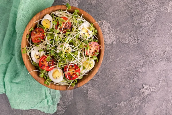 Light salad with radish sprouts, quail eggs, cherry tomatoes and sesame in wooden bowl on dark concrete background. Copy space. Top view