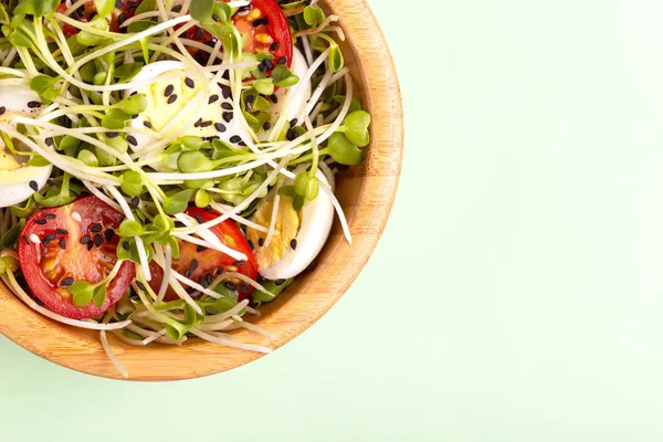 Light salad with radish sprouts, quail eggs, cherry tomatoes and sesame in wooden bowl on light green background. Copy space. Top view