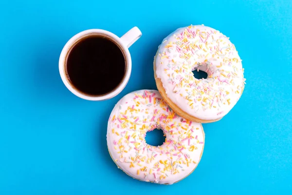 Two delicious vanilla donuts with sprinkle and cup of coffee on bright blue background. Unhealthy, but tasty sweets. Copy space. Top view