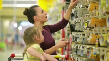 Anne ve kızı süpermarkette gülümseyerek. Bir kızın annesi bir yapışkan bant seçin yardımcı olur. Tatlı kızı bir arabası içinde oturuyor. Çok birlikte eğlenceli yaşıyorsanız.