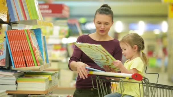 Ung mamma med hennes dotter välja böcker i snabbköpet. Vackra dotter sitter i en stormarknad vagn och anser illustrationerna. — Stockvideo