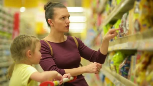Madre e hija Compras en el supermercado. Están comprando copos de desayuno. Una hija sentada en un carro de supermercado . — Vídeos de Stock