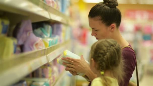 Cutie Girl con lecca-lecca seduto nel carrello del supermercato. Madre sullo sfondo sceglie salviettine umidificate . — Video Stock