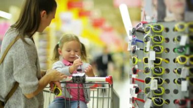 Genç karısı Casual giyim güneş gözlüğü için küçük kızı ayakta süpermarket raf seçme.
