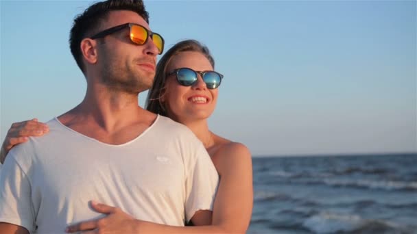 Vista frontal de la feliz pareja sonriente con gafas de sol de moda abrazándose en la playa cerca del mar durante el soleado día de verano . — Vídeo de stock