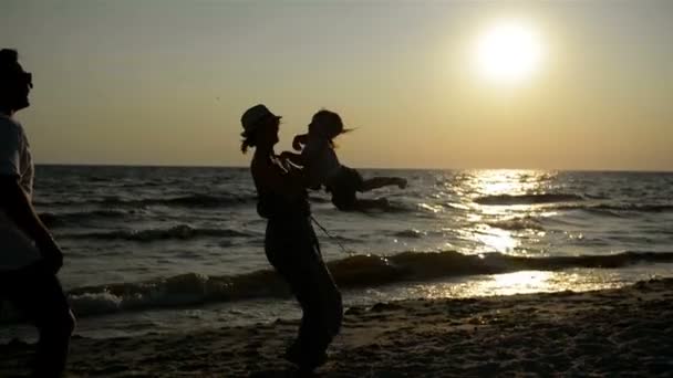 Três silhuetas escuras da mãe e do pai com a filha pequena perto do mar durante o por do sol . — Vídeo de Stock