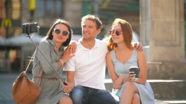 Poz ve ekşitmeden üç neşeli arkadaşları Selfie Smartphone kullanarak açık havada alarak tamamlayın. İki güzel kız ve kendilerini cep telefonuyla çekim bir yakışıklı erkek.