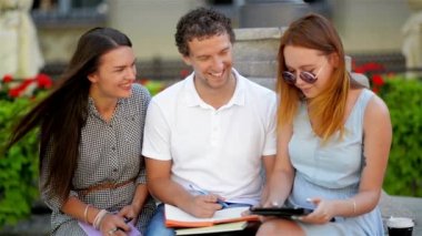 Sevimli gülümsüyor ve uzun saç oturan beyaz gömlek ve kot pantolon ve sınav kullanarak siyah Tablet, notlar ve kalemler için hazırlanıyor yakışıklı çocuk yakınındaki Brench üzerinde iki Bayan.