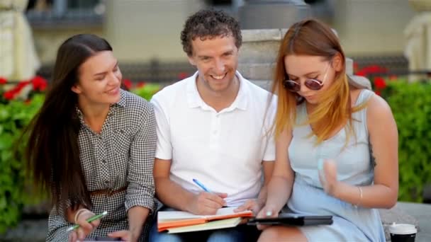 Вид спереди на трех друзей, готовящихся к совместному экзамену на открытом воздухе, сидящих на скамейке в городском парке в теплый солнечный день . — стоковое видео