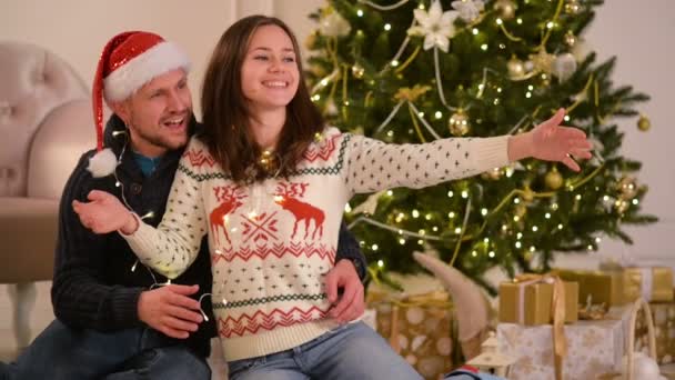 Casal jovem bonito no amor vestindo chapéus de Papai Noel, deitado no chão ao lado de uma lareira e uma árvore de Natal bem decorada e apreciando a manhã de Natal . — Vídeo de Stock