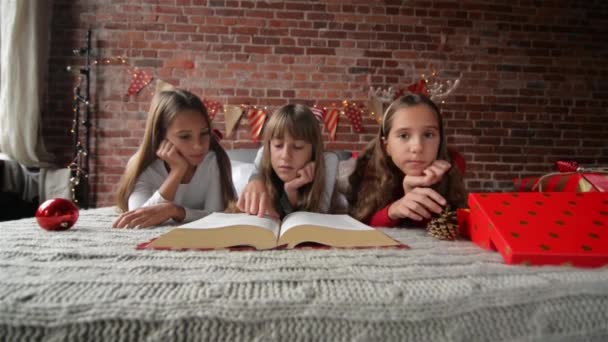 Tres hermanas yacen en la cama en pijamas suaves, trillizos leen un libro enorme de historias de Navidad, el dormitorio está decorado con luces y regalos de Navidad — Vídeos de Stock