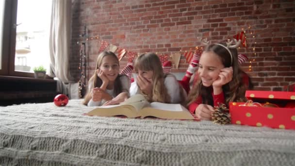 Tres hermanas yacen en la cama en pijamas suaves, trillizos leen un libro enorme de historias de Navidad, el dormitorio está decorado con luces y regalos de Navidad — Vídeos de Stock