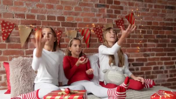 Tres hermanas sentadas en la cama en pijamas suaves y jugando con manzanas rojas. El dormitorio está decorado con luces de Navidad. Feliz Navidad y un feliz año nuevo . — Vídeos de Stock