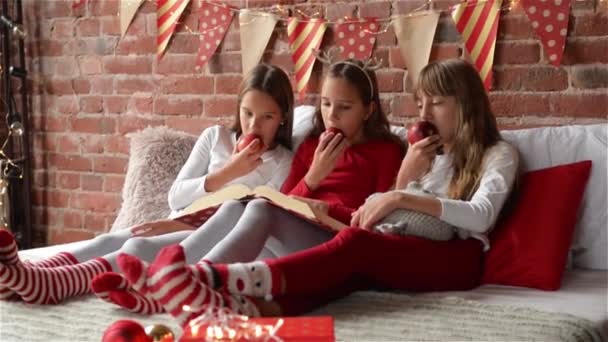Три сестры сидят на кровати в рождественской пижаме и едят красные яблоки, Триплеты девочки читают книгу рождественских историй, спальня украшена рождественскими огнями и подарками — стоковое видео