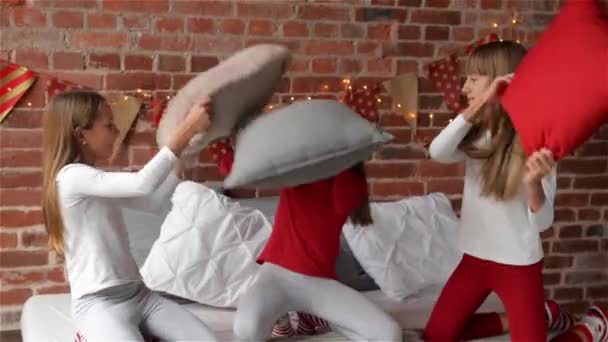 Drillinge Schwestern im Weihnachtsschlafanzug haben eine Kissenschlacht auf dem Bett, das für Weihnachten, frohe Weihnachten und ein gutes neues Jahr dekoriert ist — Stockvideo